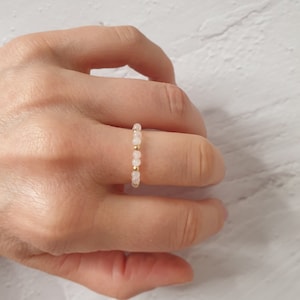 May include: A delicate ring made of small white beads and gold accents. The ring is worn on a finger.