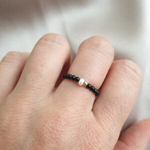 May include: A beaded ring featuring small black beads, a single white bead, and a gold-colored bead. The ring is worn on a finger, showcasing its delicate design and contrasting colors. The background is a soft, white fabric.
