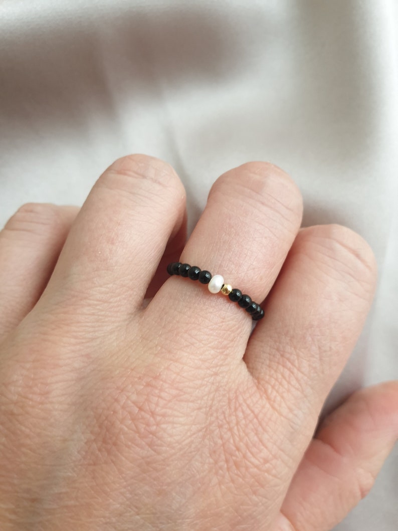 May include: A beaded ring featuring small black beads, a single white bead, and a gold-colored bead. The ring is worn on a finger, showcasing its delicate design and contrasting colors. The background is a soft, white fabric.