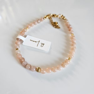 K&ouml;nnte beinhalten: Ein zartes Armband aus hellrosa Perlen mit goldenen Akzenten. Das Armband hat ein kleines Etikett mit dem Text "8 mm" befestigt.