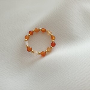 May include: A beaded ring featuring a circular design. The ring is composed of small, round beads in shades of orange, white, and gold. The beads are strung together with gold-colored spacers. The ring is displayed on a white, textured surface.