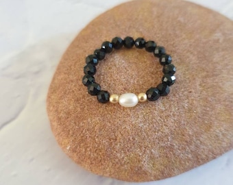 May include: A black beaded ring with a central white pearl and gold accents. The ring is composed of small, faceted black beads, creating a sparkling effect. The pearl is oval-shaped and sits between two gold beads.