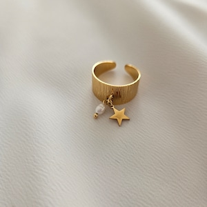 May include: Gold-colored adjustable ring with a textured band. A small gold star and a white pearl dangle from the ring. The ring is set against a white, textured background.