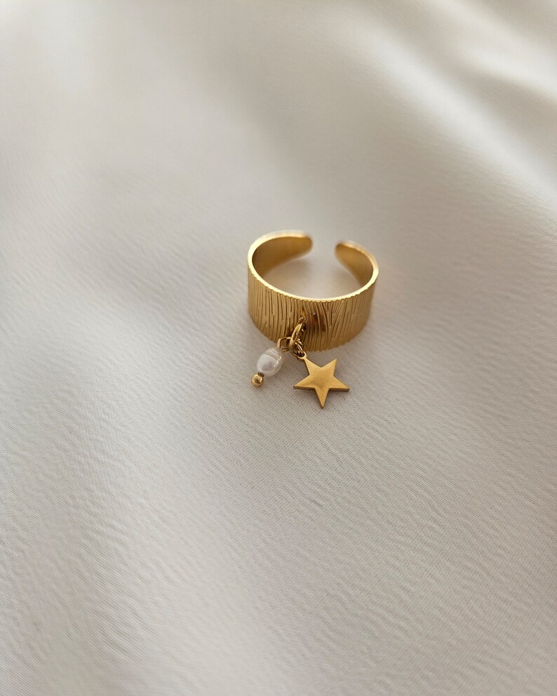 May include: Gold-colored adjustable ring with a textured band. A small gold star and a white pearl dangle from the ring. The ring is set against a white, textured background.