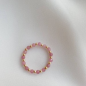 May include: A delicate ring made of pink beads and gold accents. The ring is made of small, round beads that are strung together to form a circle.