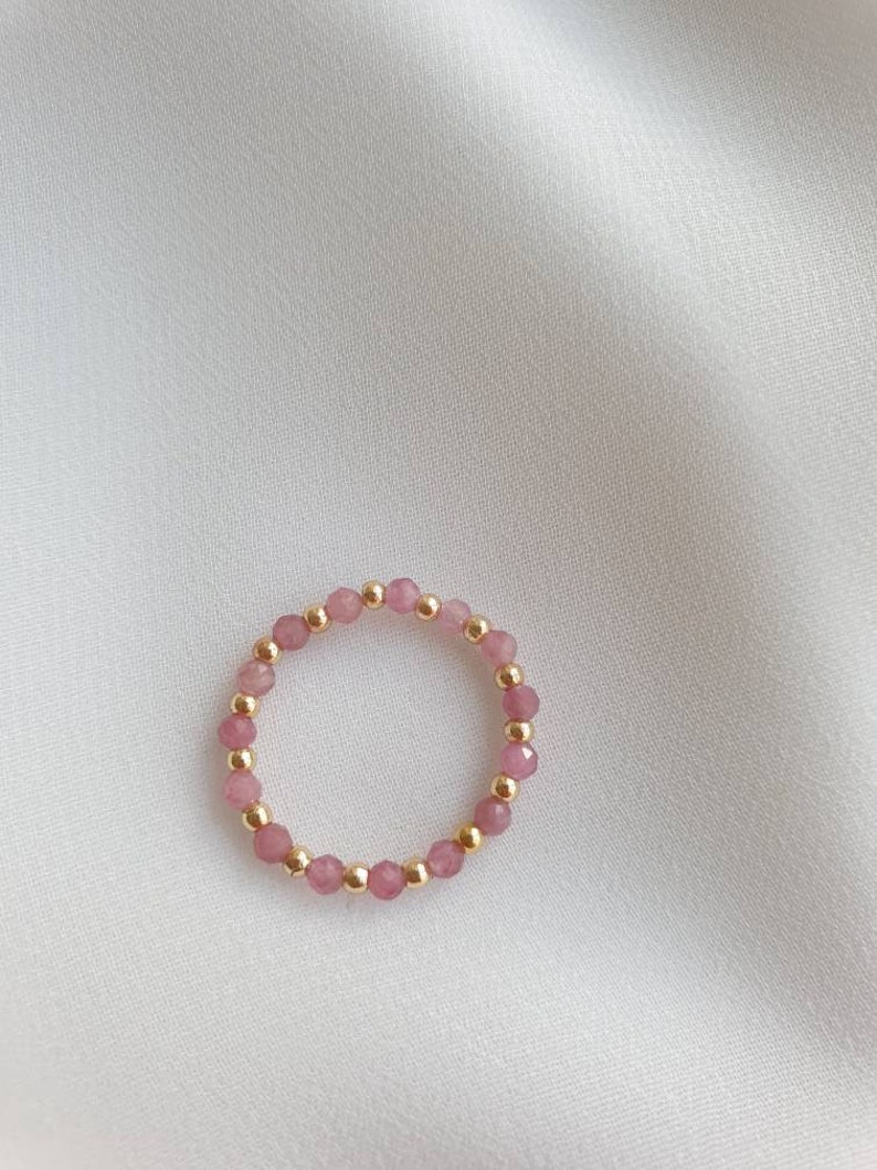 May include: A delicate ring made of pink beads and gold accents. The ring is made of small, round beads that are strung together to form a circle.