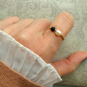 May include: A gold-colored ring featuring a black bead and a white pearl. The ring is worn on a finger, with a white and tan fabric in the background. The ring is a delicate piece of jewelry.