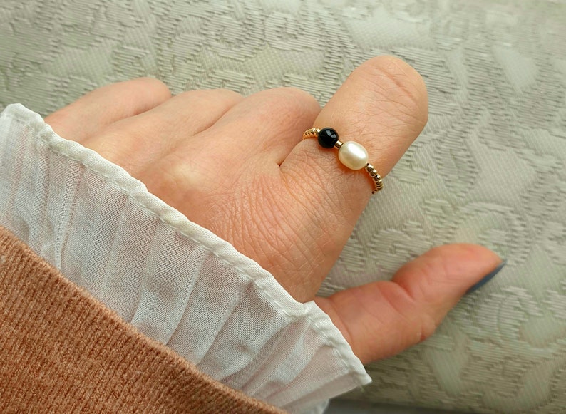 May include: A gold-colored ring featuring a black bead and a white pearl. The ring is worn on a finger, with a white and tan fabric in the background. The ring is a delicate piece of jewelry.