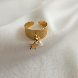 May include: Gold-toned adjustable ring with a textured band. A small gold star and a white pearl charm dangle from the ring. The ring is set against a white, textured background.