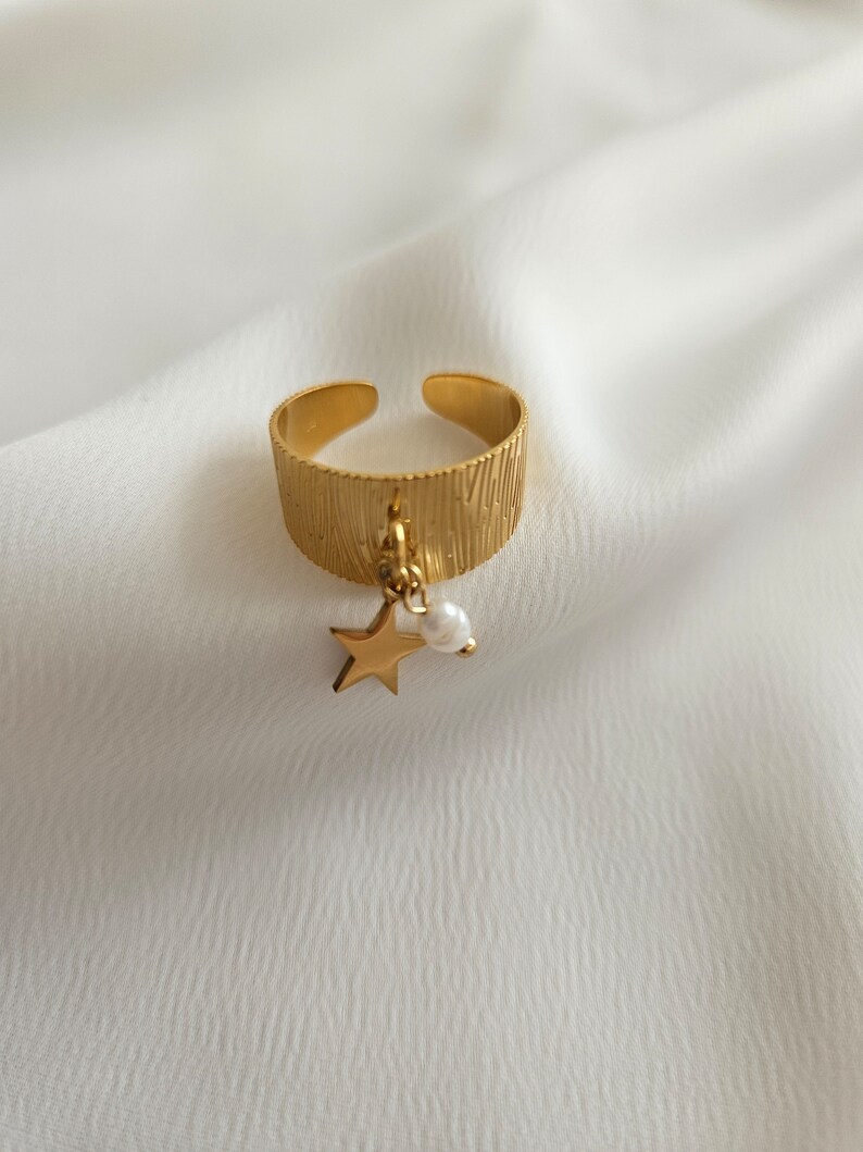 May include: Gold-toned adjustable ring with a textured band. A small gold star and a white pearl charm dangle from the ring. The ring is set against a white, textured background.