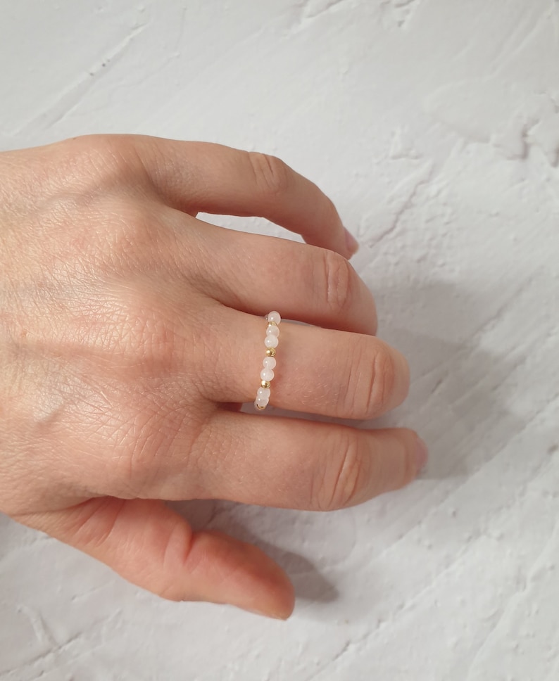 May include: A delicate ring made of small pink beads and gold spacers. The ring is worn on a finger.
