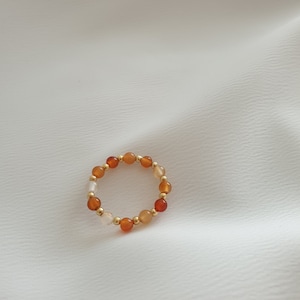 May include: A beaded ring featuring a mix of orange, red, and cream-colored gemstones, interspersed with small gold-colored beads. The ring is set against a white, textured background, showcasing its colors and design.