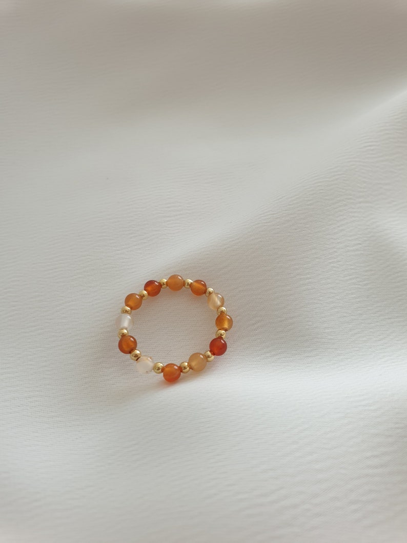 May include: A beaded ring featuring a mix of orange, red, and cream-colored gemstones, interspersed with small gold-colored beads. The ring is set against a white, textured background, showcasing its colors and design.