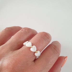 Pode incluir: Um anel branco com contas em forma de coração e pequenas contas douradas. O anel é usado num dedo. O fundo é branco liso.