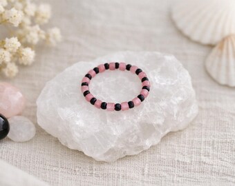 May include: A beaded ring featuring alternating pink and black beads. The ring is displayed on a white, textured surface, with other decorative elements in the background, including seashells and gemstones.