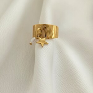 May include: Gold-colored ring with a textured surface and a dangling star charm. The ring features a small star pendant and a white pearl-like bead. The ring is displayed on a white fabric background.