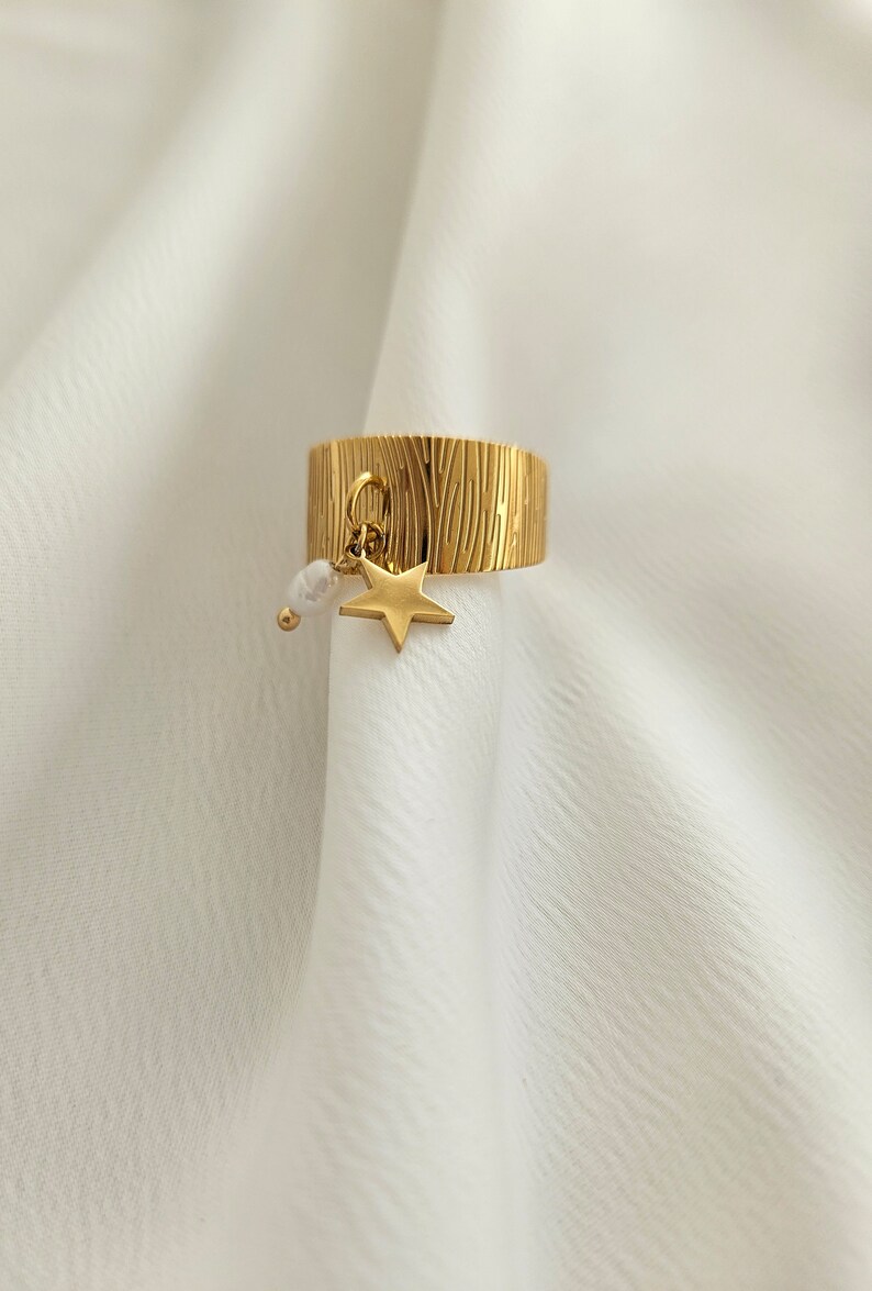 May include: Gold-colored ring with a textured surface and a dangling star charm. The ring features a small star pendant and a white pearl-like bead. The ring is displayed on a white fabric background.