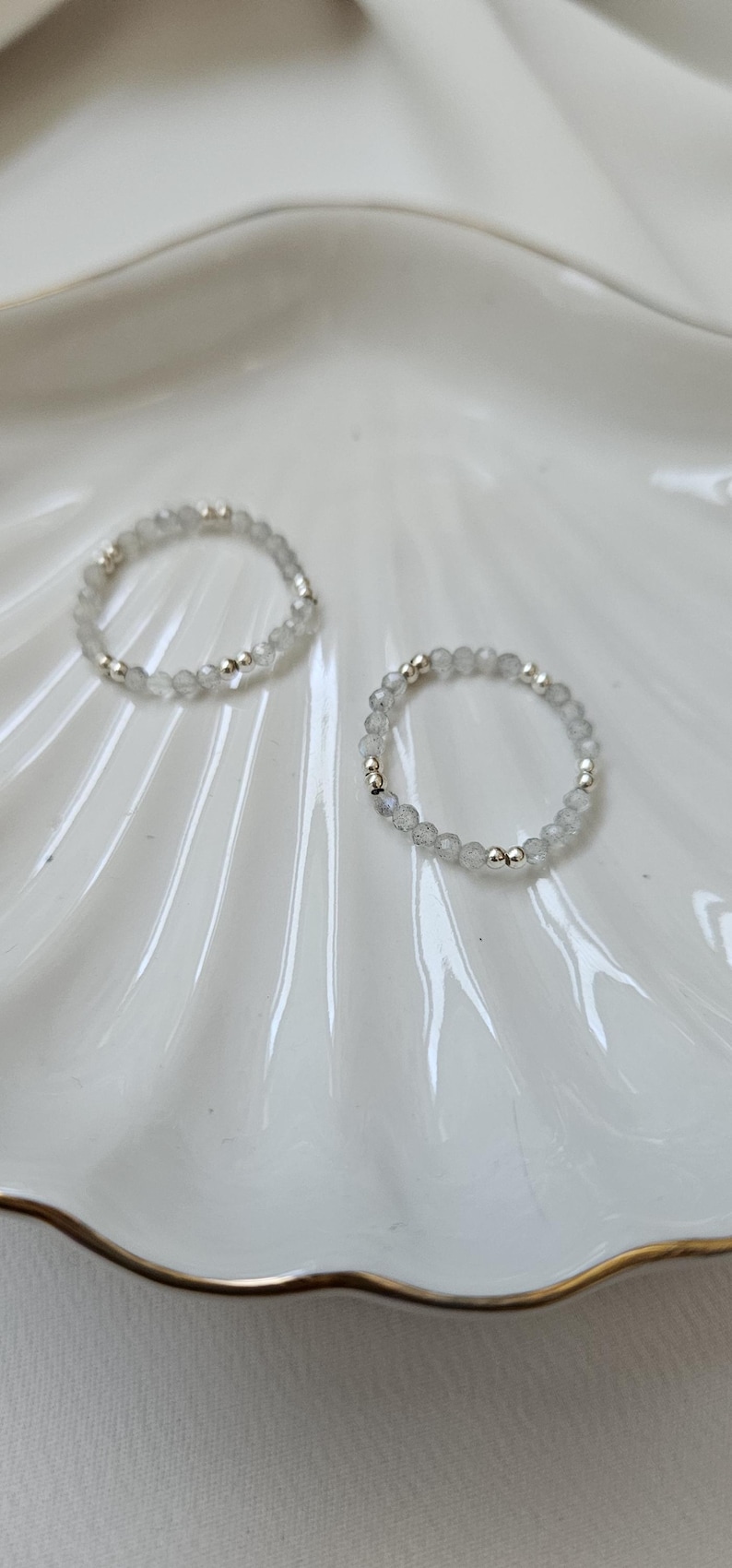 May include: Two silver rings with white gemstone beads on a white ceramic plate with a gold rim.