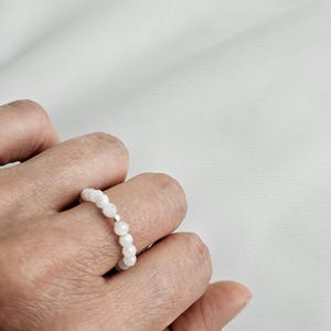 May include: A white beaded ring is worn on a finger. The ring is made of small, round, white beads strung together. A small silver-colored spacer is visible between the beads. The ring is on a person's finger.