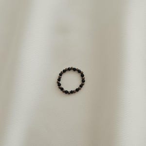 K&ouml;nnte beinhalten: Ein schwarzer und goldener Perlenring. Der Ring besteht aus kleinen, runden Perlen, die auf einem d&uuml;nnen Draht aufgereiht sind.