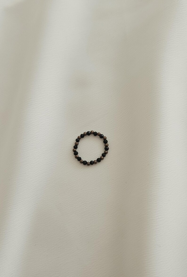 K&ouml;nnte beinhalten: Ein schwarzer und goldener Perlenring. Der Ring besteht aus kleinen, runden Perlen, die auf einem d&uuml;nnen Draht aufgereiht sind.