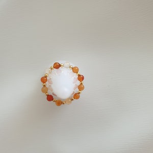 May include: A beaded ring featuring orange and white gemstone beads interspersed with small gold-colored beads. The ring is set against a white background, showcasing the jewelry's colors and design.