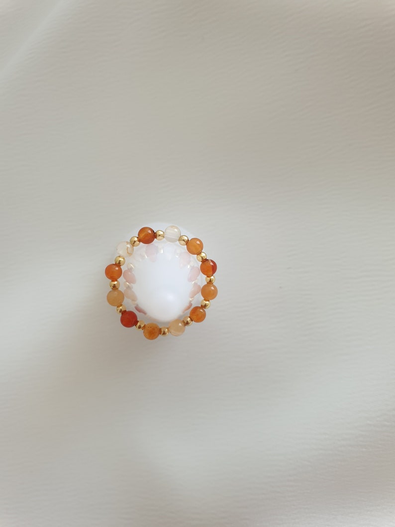 May include: A beaded ring featuring orange and white gemstone beads interspersed with small gold-colored beads. The ring is set against a white background, showcasing the jewelry's colors and design.
