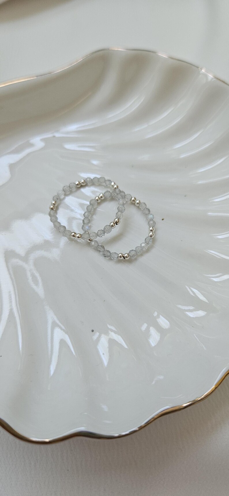 May include: Two silver beaded bracelets with clear faceted stones. The bracelets are on a white ceramic plate with a gold rim.