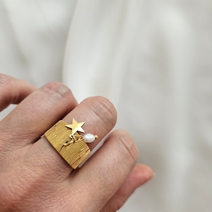 May include: A gold-colored ring with a textured, rectangular face. A small gold star and a dangling white pearl are attached to the ring. The ring is worn on a finger.