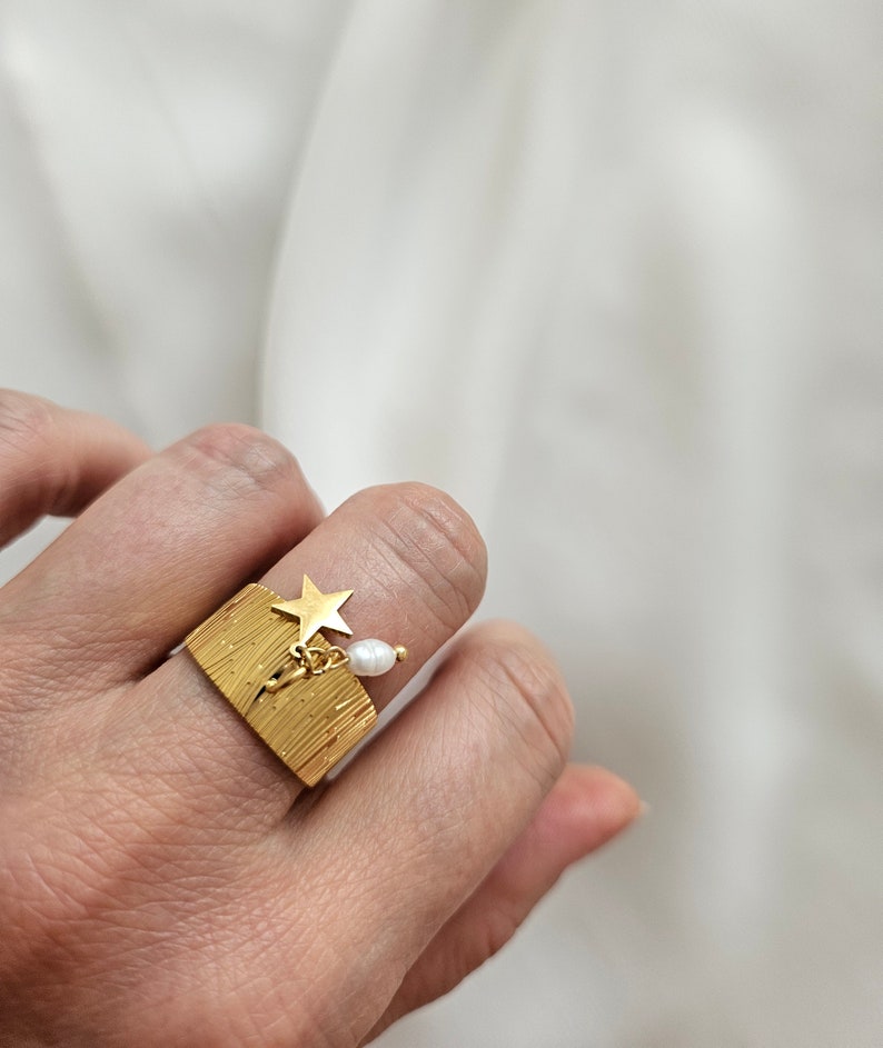 May include: A gold-colored ring with a textured, rectangular face. A small gold star and a dangling white pearl are attached to the ring. The ring is worn on a finger.