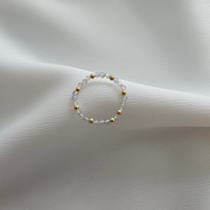 May include: A delicate ring made of clear faceted beads and gold accents. The ring is on a white fabric background.