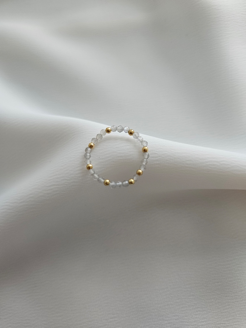 May include: A delicate ring made of clear faceted beads and gold accents. The ring is on a white fabric background.