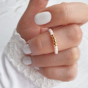 May include: A beaded ring featuring small white and gold beads. The ring is worn on a finger, showcasing the delicate design and color contrast. The gold beads are clustered together, adding a touch of elegance to the simple design.
