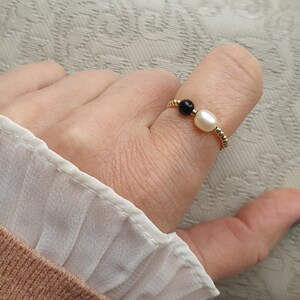 May include: A gold-toned ring featuring a white pearl and a black bead. The ring is worn on a finger, showcasing its delicate design. The ring's band has a textured, beaded appearance, adding to its elegance. The ring is a piece of jewelry.