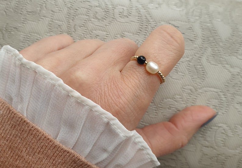 May include: A gold-toned ring featuring a white pearl and a black bead. The ring is worn on a finger, showcasing its delicate design. The ring's band has a textured, beaded appearance, adding to its elegance. The ring is a piece of jewelry.