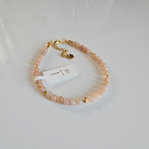 K&ouml;nnte beinhalten: Ein zartes goldenes Armband mit kleinen, hellrosa Perlen und goldenen Akzenten. Das Armband hat ein Etikett mit der Aufschrift "8 mm".