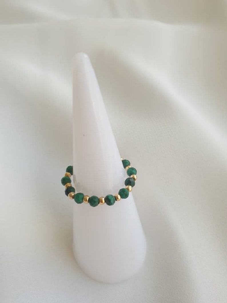 May include: A gold and green beaded ring on a white ring display stand. The ring is made of small, round beads in shades of green and gold.