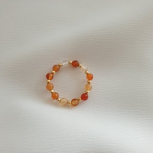May include: A beaded ring featuring a circular design. The ring is composed of small, round beads in shades of orange, white, and gold. The beads are strung together to form a continuous loop, creating a simple yet elegant piece of jewelry.