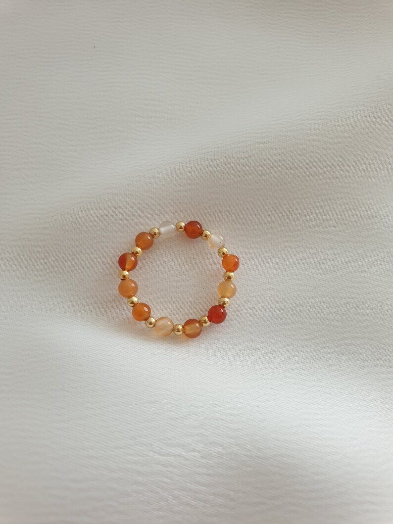 May include: A beaded ring featuring a circular design. The ring is composed of small, round beads in shades of orange, white, and gold. The beads are strung together to form a continuous loop, creating a simple yet elegant piece of jewelry.