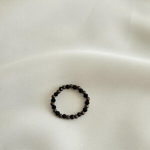 K&ouml;nnte beinhalten: Ein schwarzer und brauner Perlenring. Der Ring besteht aus kleinen, runden Perlen, die zu einem Kreis zusammengef&uuml;gt sind.