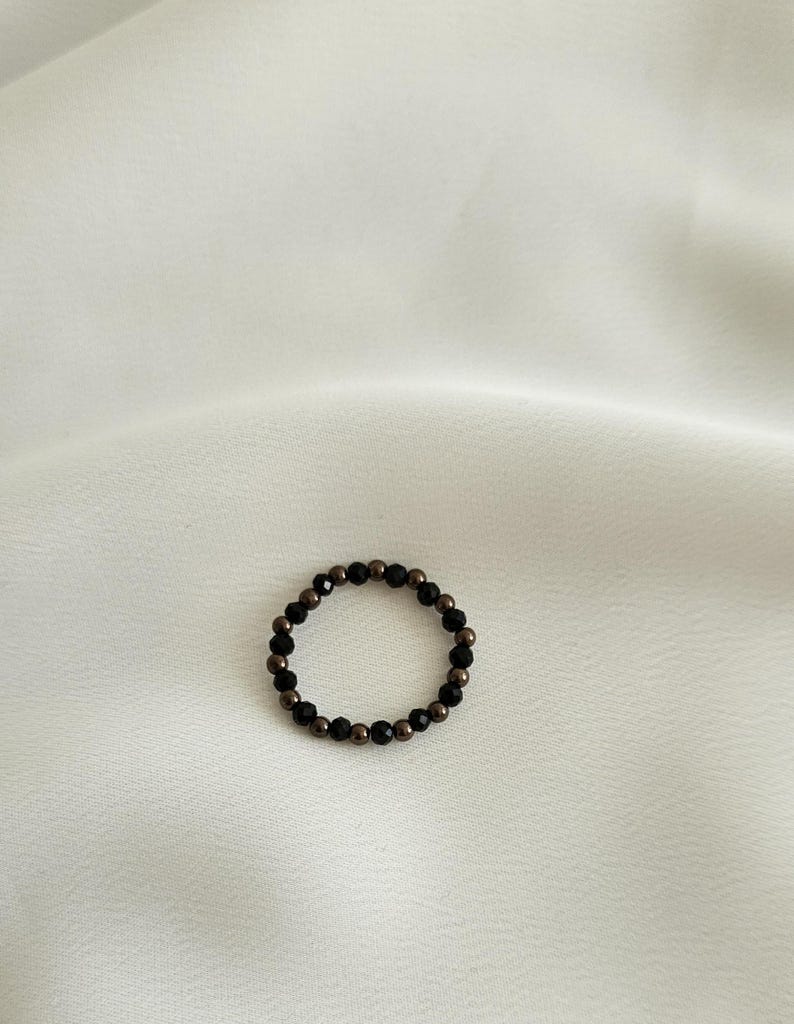 K&ouml;nnte beinhalten: Ein schwarzer und brauner Perlenring. Der Ring besteht aus kleinen, runden Perlen, die zu einem Kreis zusammengef&uuml;gt sind.