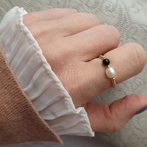 May include: A gold beaded ring featuring a white pearl and a black bead. The ring is worn on a finger, with a white ruffled sleeve and a tan sweater in the background.
