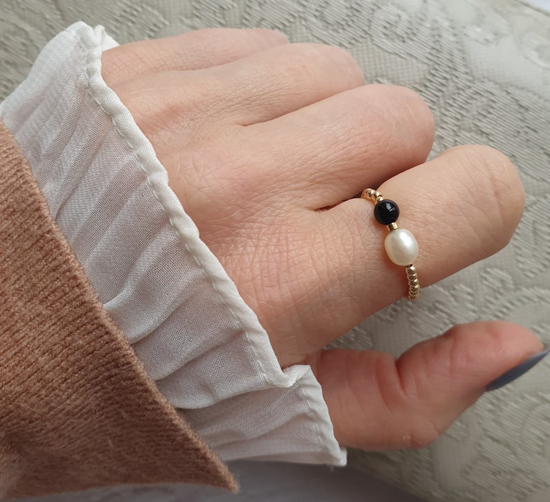 May include: A gold beaded ring featuring a white pearl and a black bead. The ring is worn on a finger, with a white ruffled sleeve and a tan sweater in the background.