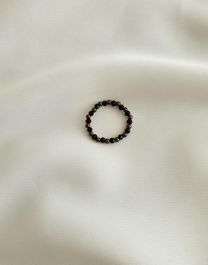 K&ouml;nnte beinhalten: Ein Ring aus schwarzen und goldenen Perlen. Der Ring besteht aus kleinen, runden Perlen, die auf einem d&uuml;nnen Draht aufgereiht sind.