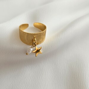 May include: Gold-colored adjustable ring with a textured band. A small gold star and a white pearl dangle from the ring. The ring is set against a white background.