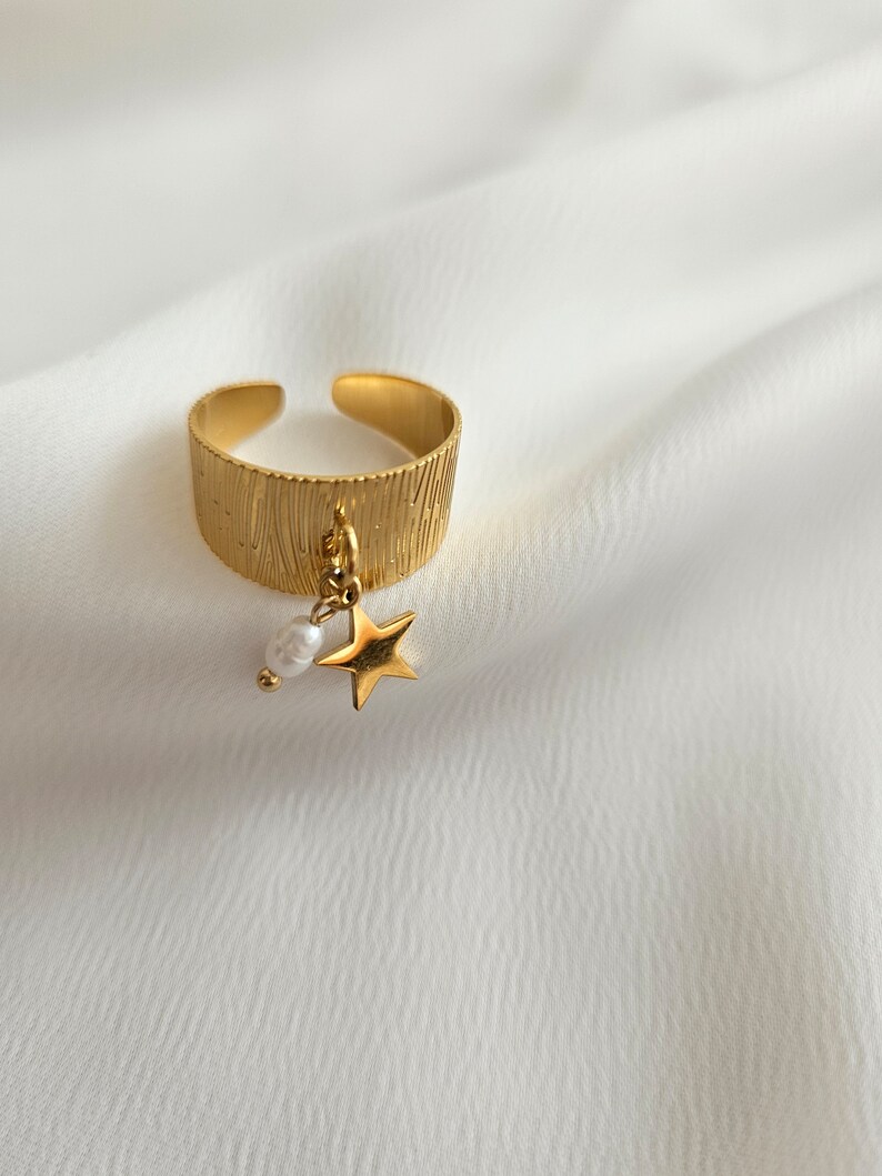 May include: Gold-colored adjustable ring with a textured band. A small gold star and a white pearl dangle from the ring. The ring is set against a white background.