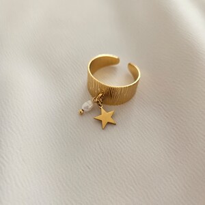 May include: Gold-colored adjustable ring with a textured band. A small gold star and a white pearl dangle from the ring. The ring is set against a white, textured background.
