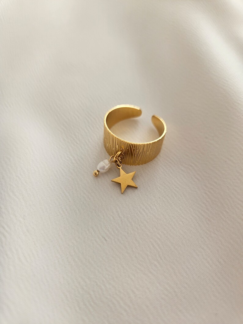 May include: Gold-colored adjustable ring with a textured band. A small gold star and a white pearl dangle from the ring. The ring is set against a white, textured background.