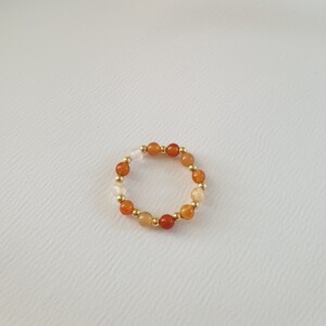 May include: A beaded ring featuring a circular design. The ring is composed of small, round beads in shades of orange, red, and cream, interspersed with gold-colored beads. The ring is set against a plain white background.
