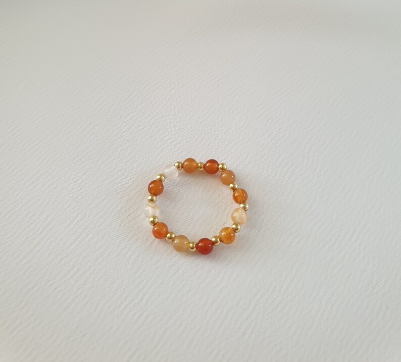 May include: A beaded ring featuring a circular design. The ring is composed of small, round beads in shades of orange, red, and cream, interspersed with gold-colored beads. The ring is set against a plain white background.